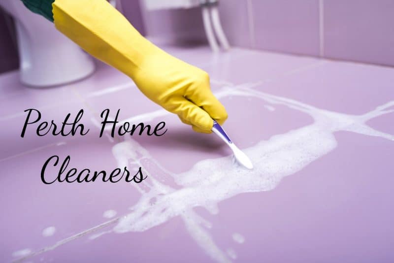 The hand of a person with a yellow glove holding a purple and white toothbrush while scrubbing the grout of a bathroom. There are soap suds where the grout has been cleaned and the base of a toilet in the background. The words Perth Home Cleaners are on the floor.