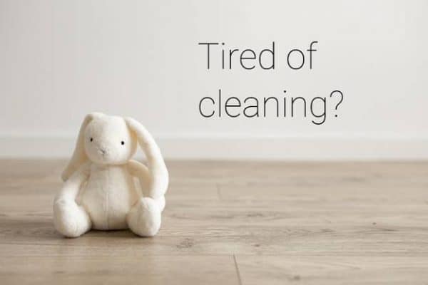 white, soft toy bunny facing the viewer, sitting on a pine floor with the words tired of cleaning over its shoulder