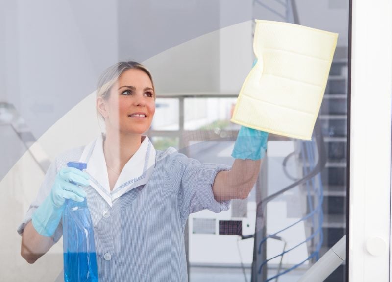 a woman holding a blue spray bottle of window cleaner and wiping a window clean